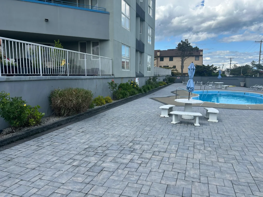 Outdoor pool area of a residential complex with gray buildings white railings and patio seating with blue umbrellas in the background