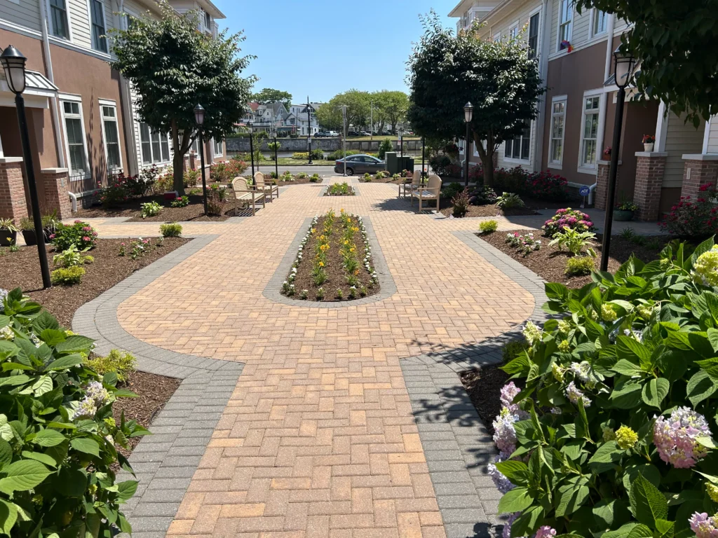 Sunlit brick paver courtyard between apartment buildings with benches flower beds and small trees street and cars visible in the background