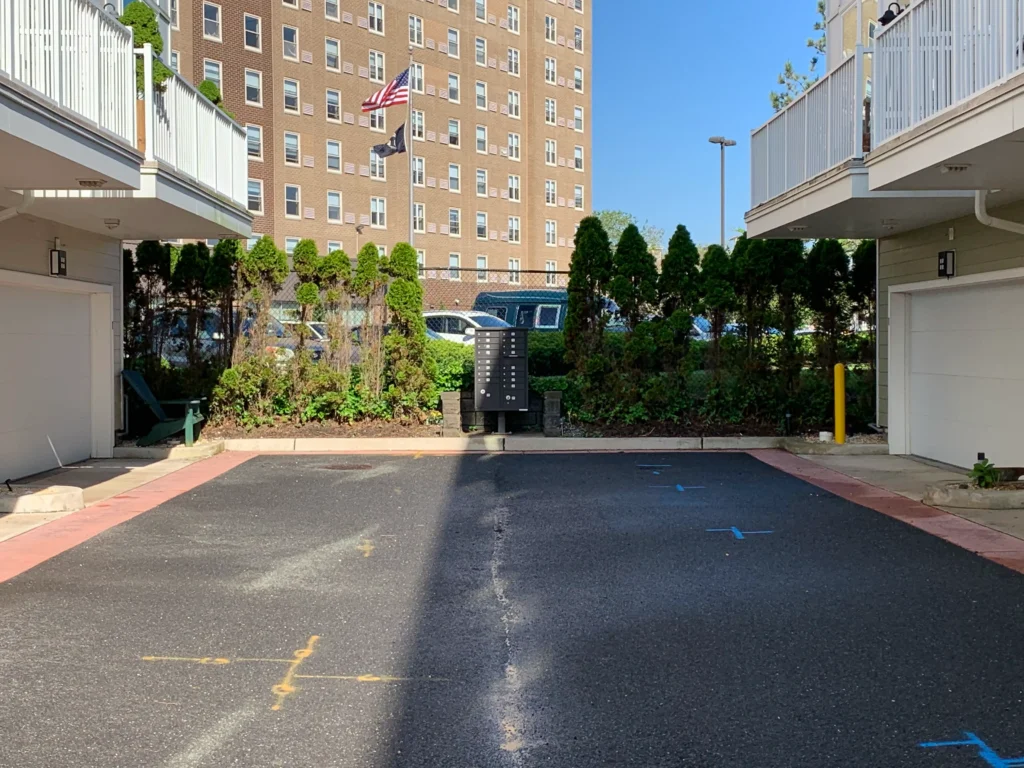 Parking lot between apartment buildings with a flagpole and American flag green shrubbery and parked cars in the background under a clear blue sky